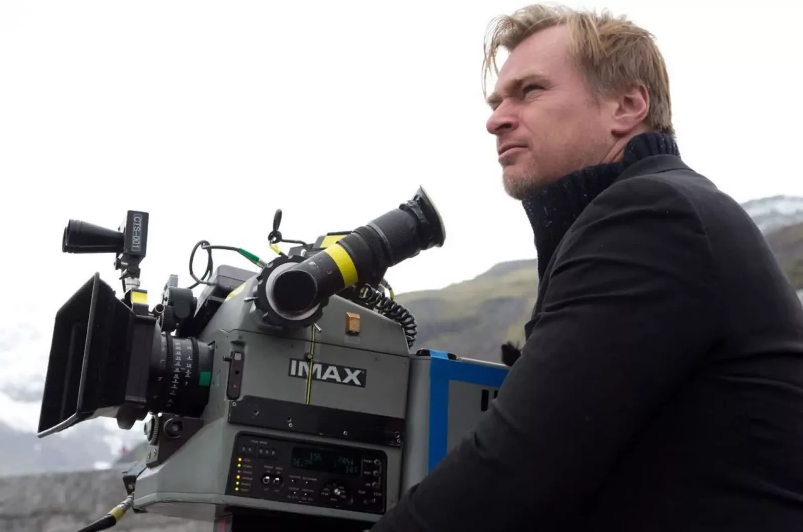Christopher Nolan looking out from behind an IMAX film camera on a mountain location shoot.