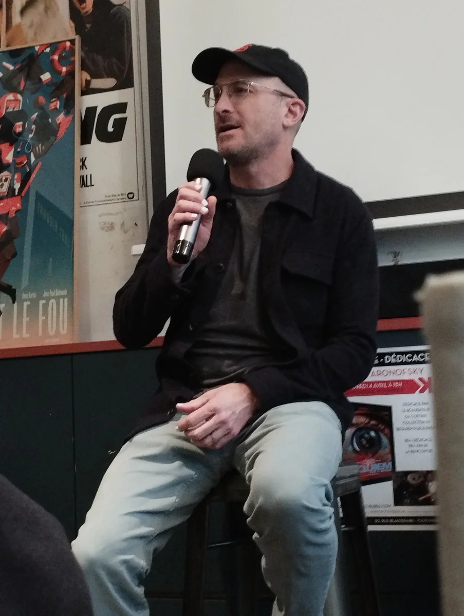 Darren Aronofsky in a cap and glasses speaking into a microphone at a film event