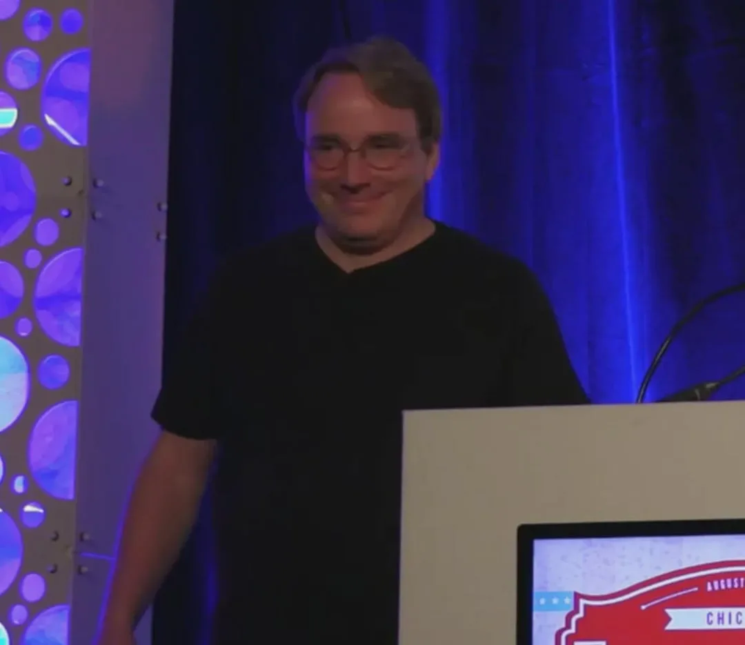 Linus Torvalds smiling at a conference podium in front of a blue-lit backdrop.