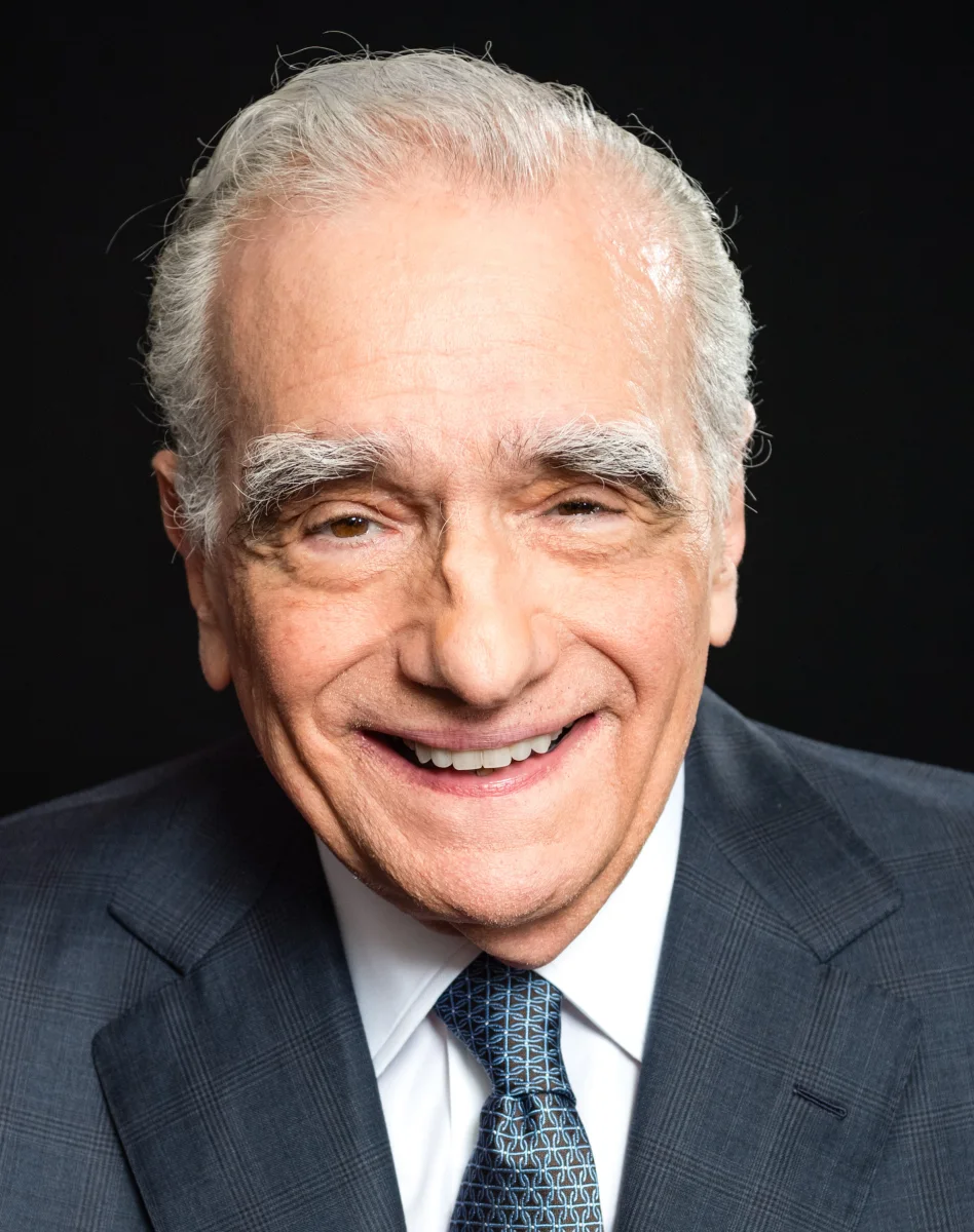 Martin Scorsese smiling in a dark plaid suit and blue tie against a black background