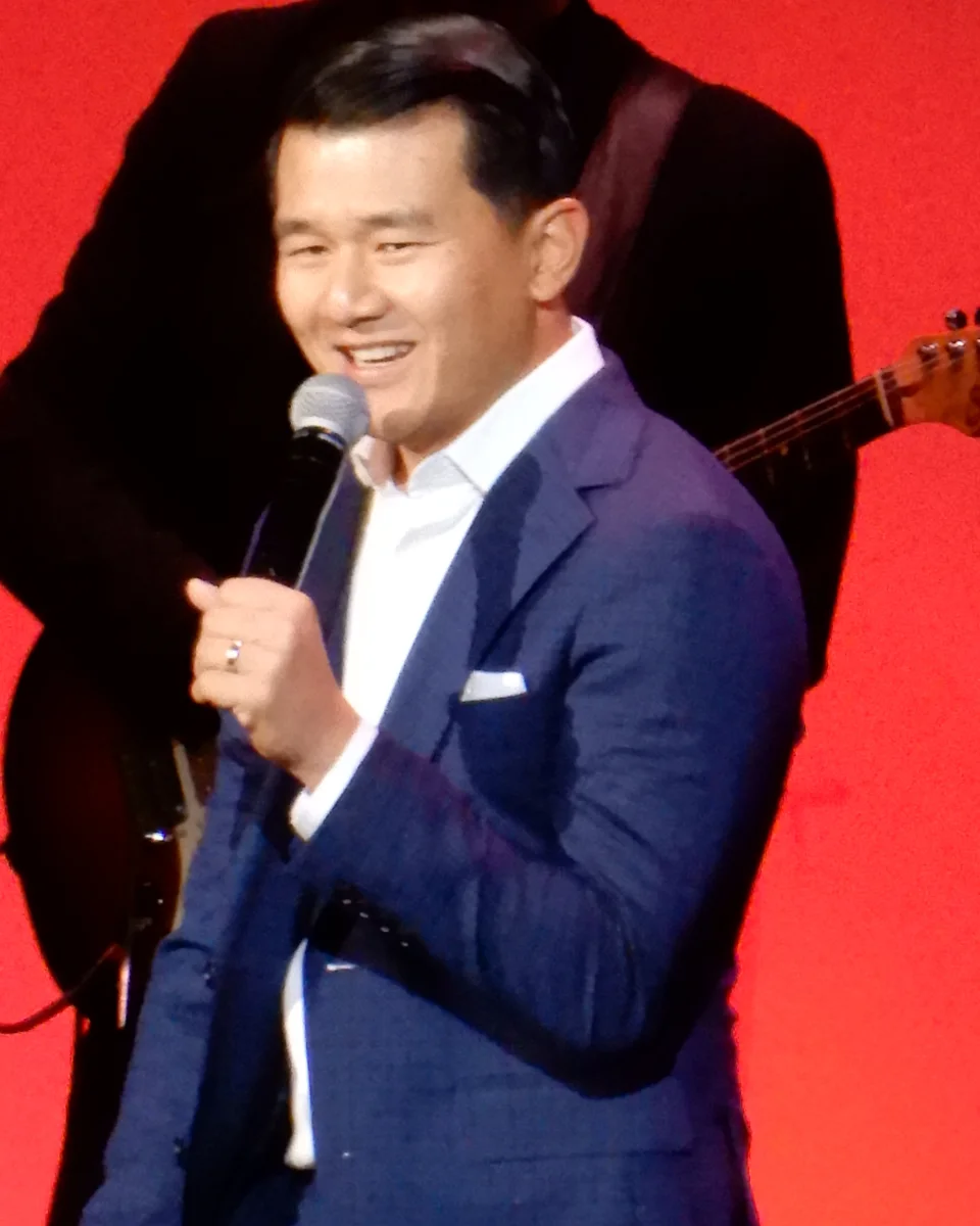 Ronny Chieng performing on stage in a blue suit at the Stand Up For Heroes benefit